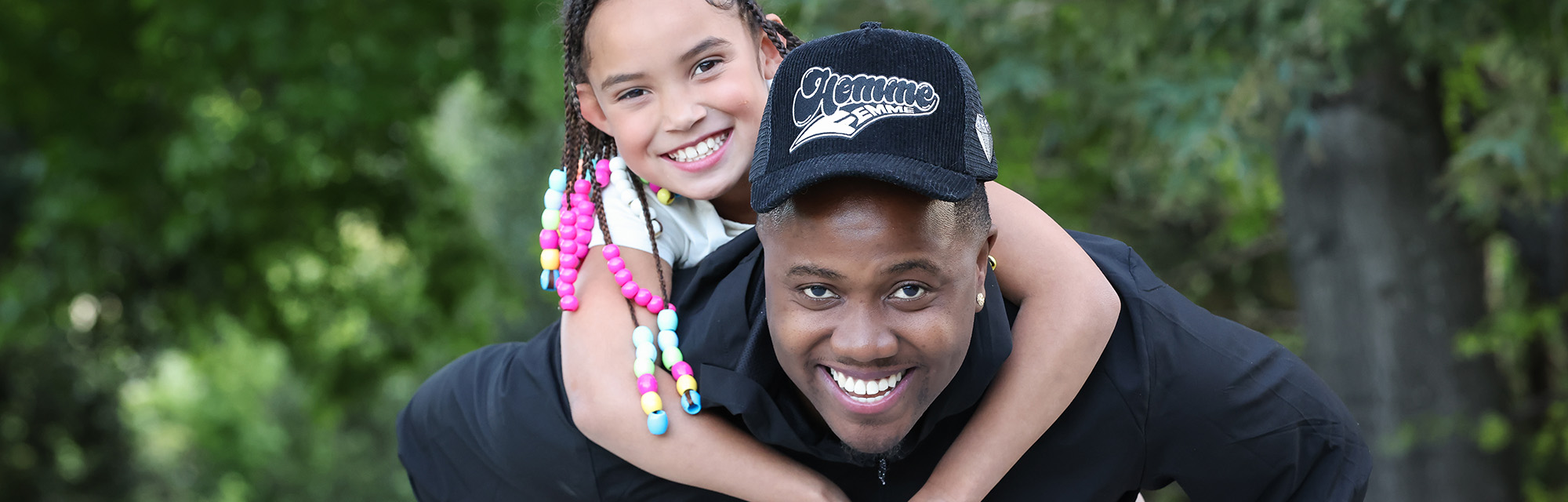Person carrying a smiling child on their back outdoors in a wooded area.