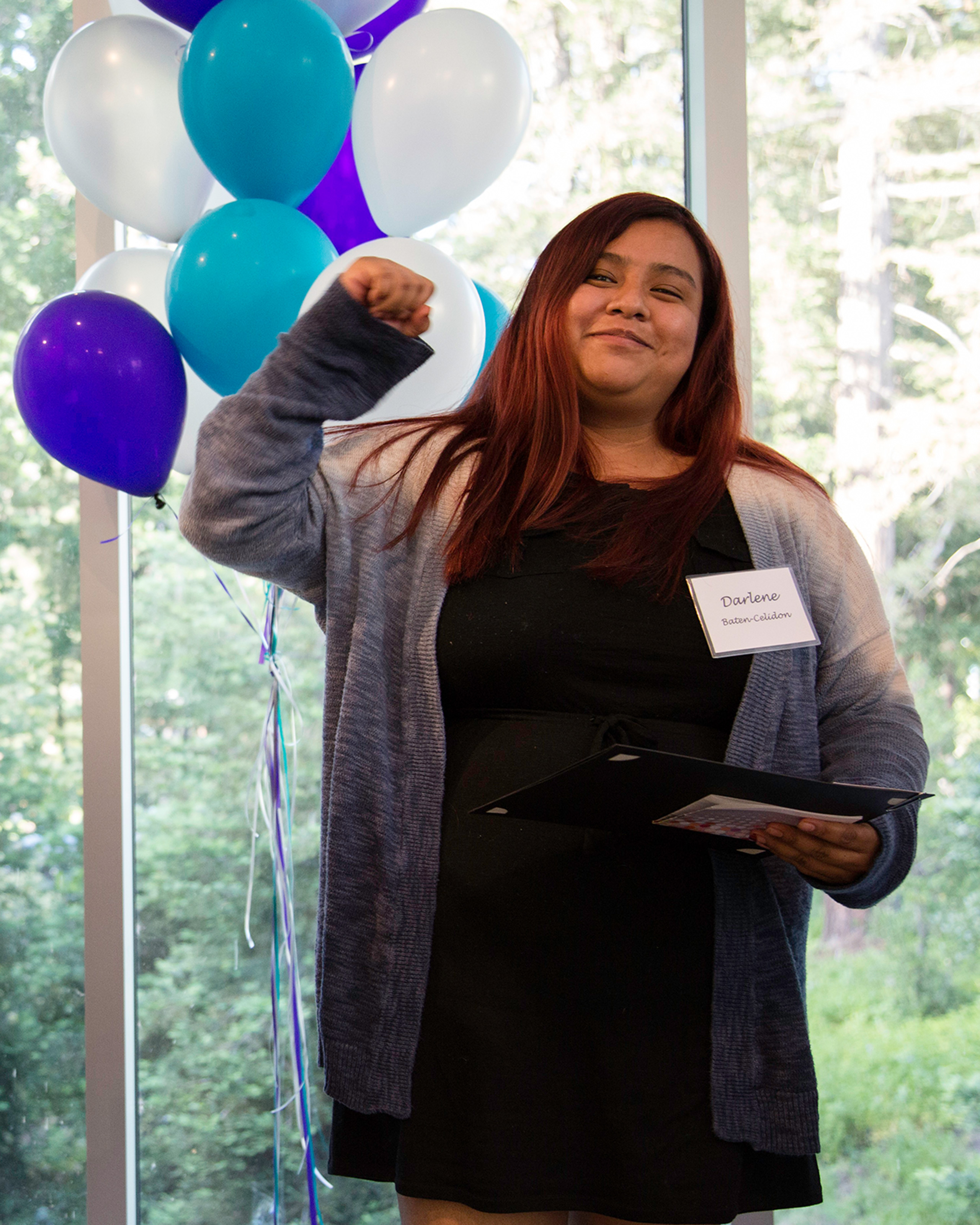 Person holding a certificate and raising a fist beside colorful balloons.
